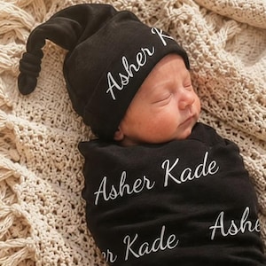 May include: A black baby swaddle blanket and matching knotted hat set featuring the name Asher Kade printed in a white cursive script repeatedly across the fabric. The swaddle wraps the infant snugly, while the hat has a knotted top detail.