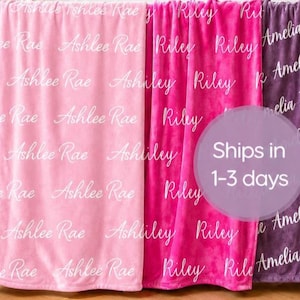 May include: Three soft blankets in light pink, hot pink, and purple, each featuring a repeating white script pattern of the names Ashlee Rae, Riley, and Amelia Jane respectively, draped over a white wooden crib frame.