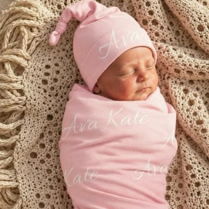 May include: A light pink baby swaddle blanket and a matching knotted beanie hat. The fabric features the name Ava Kate printed in a white cursive script repeatedly across the surface of both the swaddle and the hat, creating a repeating pattern.