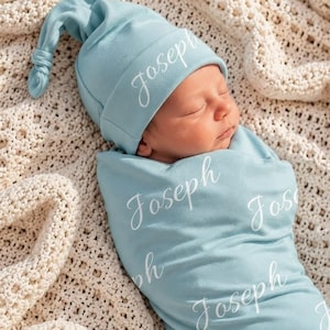 Può includere: Un set composto da una coperta per neonati e un berretto con nodo in azzurro, entrambi con il nome Joseph stampato in corsivo bianco. Il tessuto è di un colore azzurro uniforme e il set è progettato per avvolgere un neonato in sicurezza.