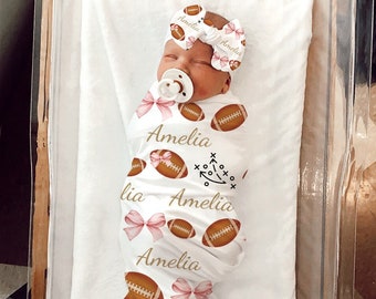 Puede incluir: Una manta de arrullo blanca con un patrón de fútbol y lazo rosa. La manta tiene el nombre "Amelia" impreso en ella varias veces. Un bebé está envuelto en la manta y lleva una diadema rosa con el nombre "Amelia" impreso en ella.