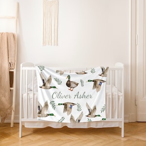 May include: White baby blanket with a green and brown duck pattern. The blanket has the name "Oliver Asher" printed in green text.
