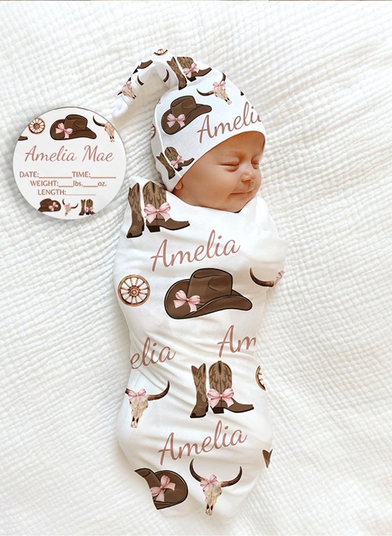 May include: A white swaddle blanket with a pink and brown cowboy boot, hat, and skull pattern. The blanket has the name "Amelia" written on it in pink. A matching pink and brown cowboy hat with the name "Amelia" written on it is on a baby's head.