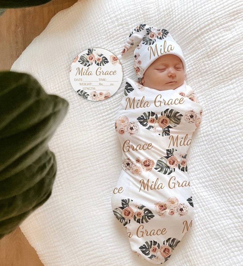 May include: A white swaddle blanket with a floral pattern and the name "Mila Grace" repeated throughout. A matching white hat with a floral pattern and the name "Mila" is on the baby's head. A round white tag with the text "Mila Grace" and spaces for date, time, weight, and length is also included.