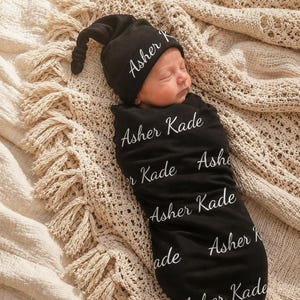 May include: A black newborn swaddle blanket and a matching knotted hat, both featuring the white cursive text Asher Kade repeated across the fabric. The swaddle is wrapped snugly around a sleeping infant, showcasing the personalized design.