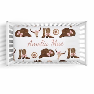 May include: White crib sheet with a brown and pink cowboy themed pattern. The sheet features cowboy hats, boots, and skulls. The name "Amelia Mae" is printed in pink on the sheet.
