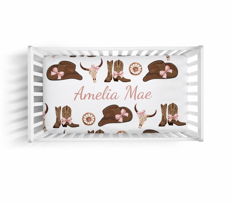 May include: White crib sheet with a brown and pink cowboy themed pattern. The sheet features cowboy hats, boots, and skulls. The name "Amelia Mae" is printed in pink on the sheet.