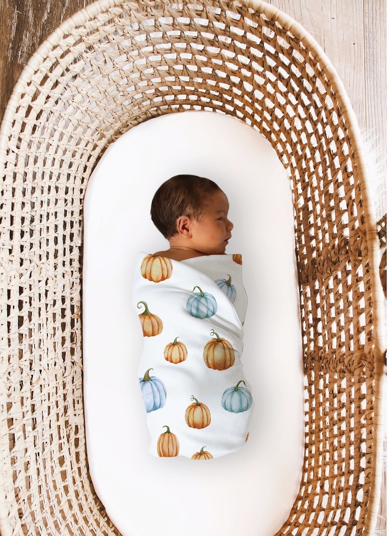 May include: A newborn baby swaddled in a white blanket adorned with watercolor pumpkins in shades of orange and blue. The baby is nestled in a woven wicker bassinet, creating a cozy and autumnal scene.