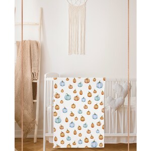 May include: White baby blanket with a repeating pattern of watercolor pumpkins in shades of orange, blue, and green. The blanket is displayed in a white crib. The brand name "gray the label" is in the top left corner.