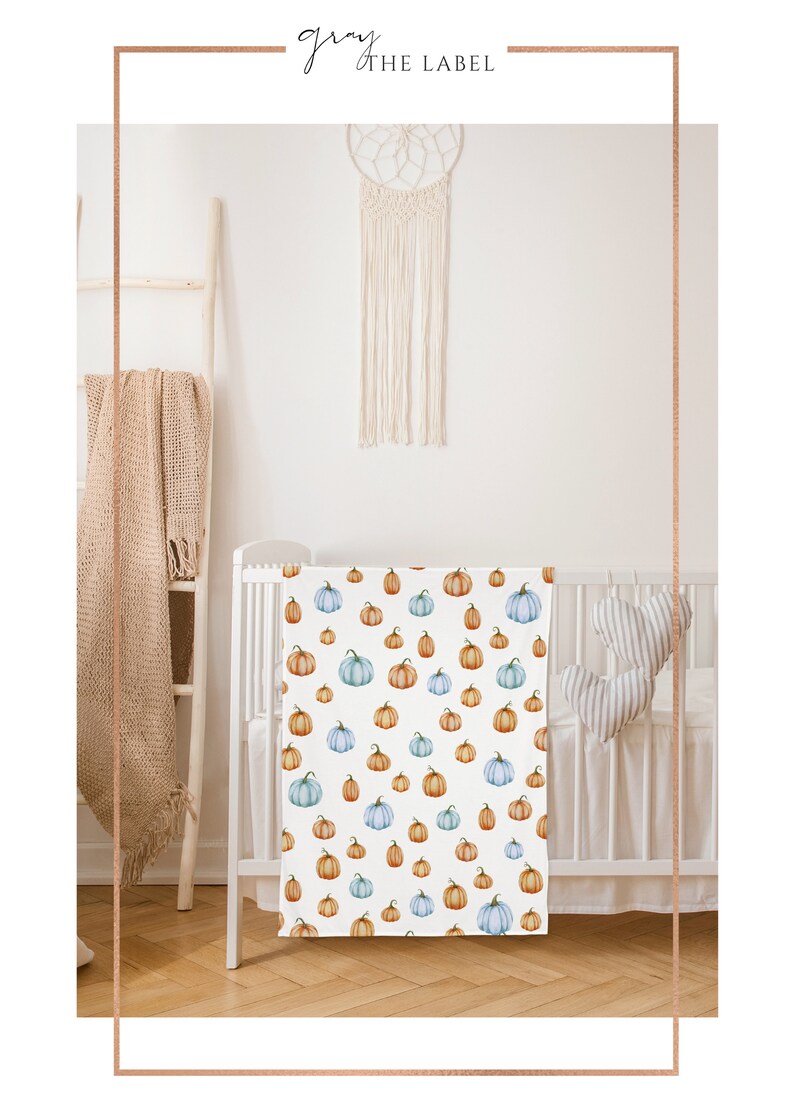May include: White baby blanket with a repeating pattern of watercolor pumpkins in shades of orange, blue, and green. The blanket is displayed in a white crib. The brand name "gray the label" is in the top left corner.