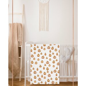 May include: A white crib with a white and orange pumpkin patterned crib sheet. The crib is in a room with a white wall and a macrame dream catcher hanging above it.