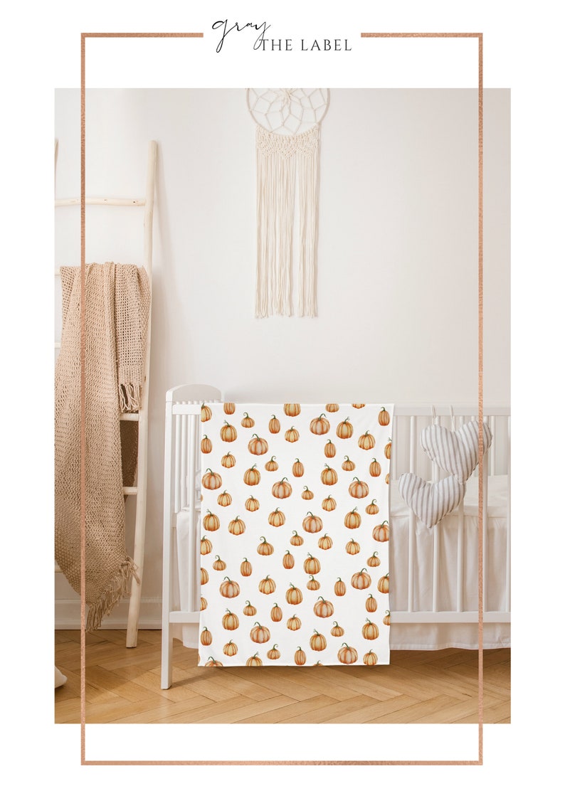 May include: A white crib with a white and orange pumpkin patterned crib sheet. The crib is in a room with a white wall and a macrame dream catcher hanging above it.
