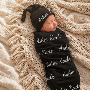 May include: A black newborn swaddle blanket and a matching knotted hat, both featuring the white printed name Asher Kade in a cursive script. The fabric is black with the name repeated multiple times across the surface of the swaddle and hat.