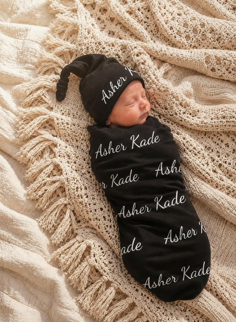 May include: A black newborn swaddle blanket and a matching knotted hat, both featuring the white printed name Asher Kade in a cursive script. The fabric is black with the name repeated multiple times across the surface of the swaddle and hat.