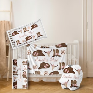 May include: A white crib bedding set with a pink and brown cowboy theme. The set includes a fitted sheet, a crib blanket, a pillowcase, and a car seat cover. The bedding features a pattern of cowboy hats, boots, and skulls. The name "Amelia Mae" is printed on each piece of bedding.