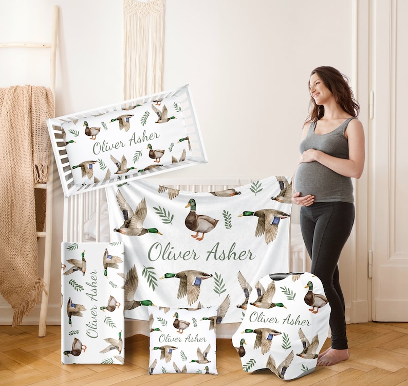 May include: A nursery bedding set with a white background and a pattern of green leaves and brown ducks. The set includes a crib sheet, a blanket, a pillow, and a car seat cover. The name "Oliver Asher" is printed on each piece.