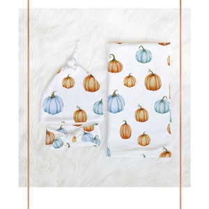 May include: A white baby set featuring a knotted hat, headband, and blanket, all adorned with watercolor pumpkins in shades of orange and blue. The set is designed for newborns and infants.