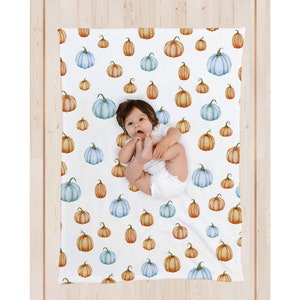 May include: White baby blanket with a pattern of watercolor pumpkins in shades of orange, blue, and green. The blanket features the text "gray the label" in a script font. A baby is lying on the blanket.