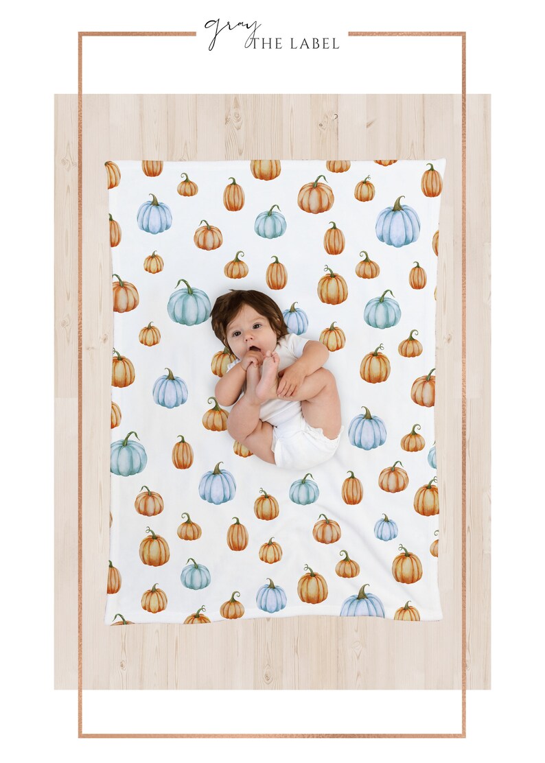 May include: White baby blanket with a pattern of watercolor pumpkins in shades of orange, blue, and green. The blanket features the text "gray the label" in a script font. A baby is lying on the blanket.