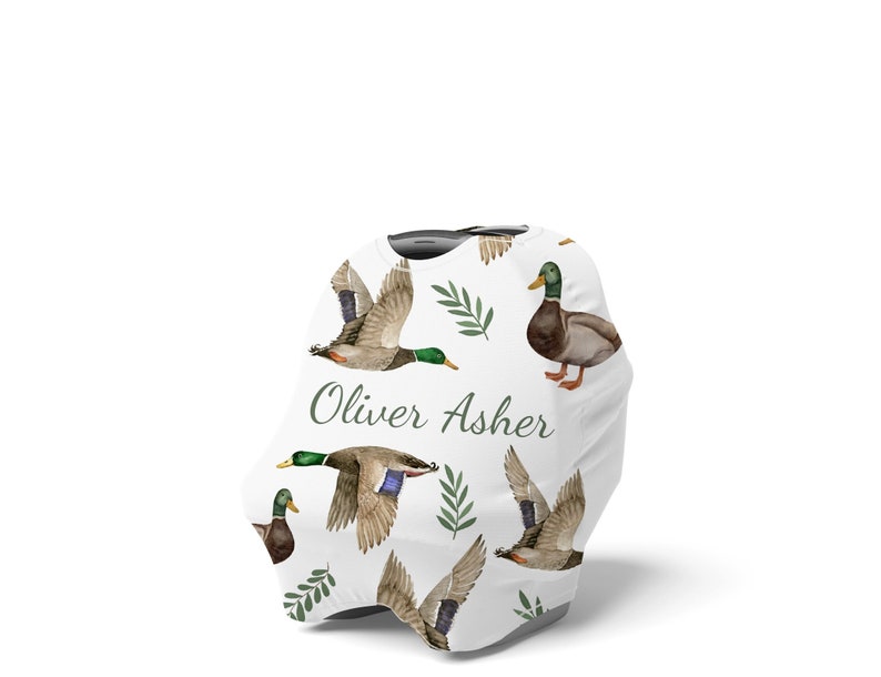 May include: White car seat cover with a pattern of green leaves and brown and green ducks. The text "Oliver Asher" is printed in green on the cover.