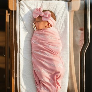 May include: A newborn infant wrapped in a soft pink swaddle blanket featuring the repeated white script text Amelia Mae. The infant wears a matching pink headband with a large bow and a white pacifier, resting peacefully in a clear bassinet.
