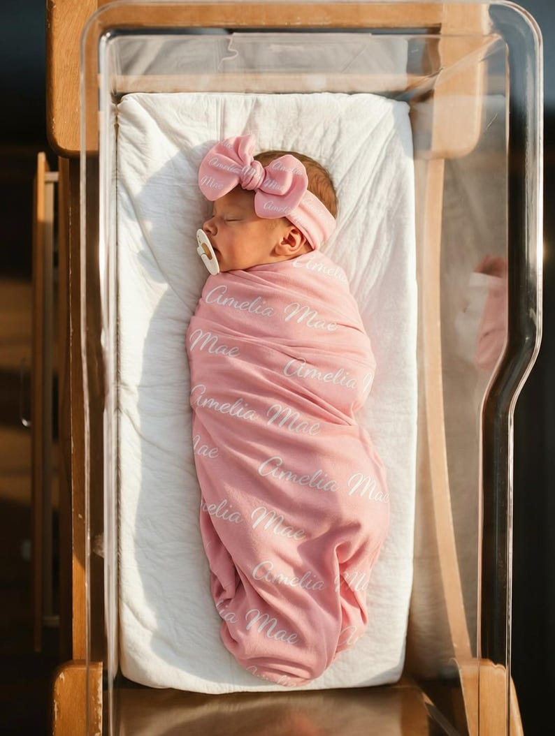 May include: A newborn infant wrapped in a soft pink swaddle blanket featuring the repeated white script text Amelia Mae. The infant wears a matching pink headband with a large bow and a white pacifier, resting peacefully in a clear bassinet.