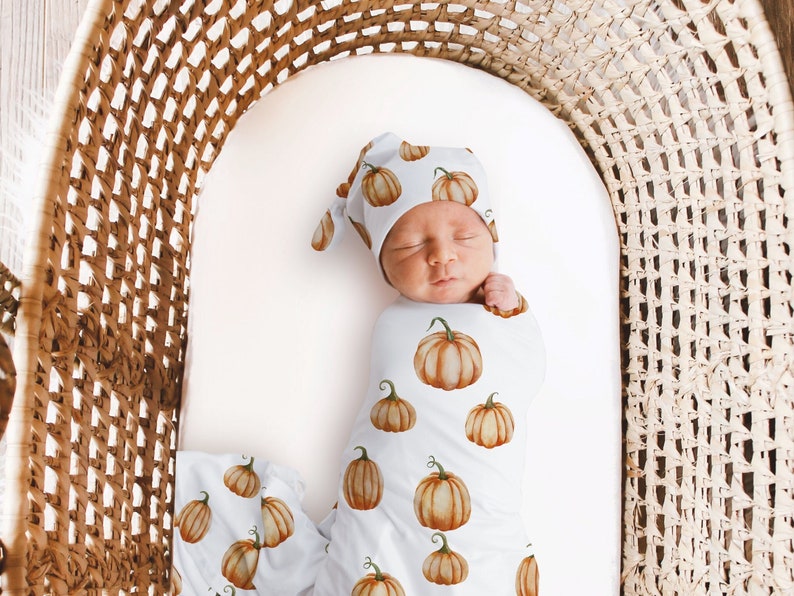 May include: A baby swaddled in a white swaddle with a pumpkin print. The baby is wearing a matching pumpkin hat. The baby is lying in a wicker basket.