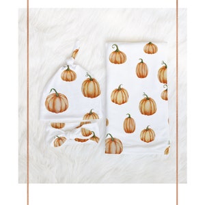 May include: A set of two baby items featuring a pumpkin print. The set includes a knotted hat and a headband. Both items are white with orange pumpkins.