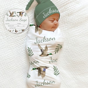 May include: A white swaddle blanket with a green and brown duck print. The blanket has the name "Jackson" written on it multiple times. A green hat with the name "Jackson" embroidered on it is on top of the blanket. A round wooden tag with the name "Jackson Sage" and spaces for birth information is also included.