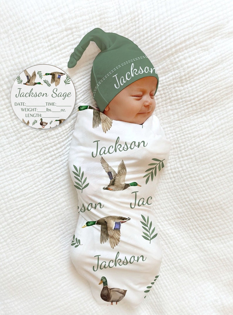 May include: A white swaddle blanket with a green and brown duck print. The blanket has the name "Jackson" written on it multiple times. A green hat with the name "Jackson" embroidered on it is on top of the blanket. A round wooden tag with the name "Jackson Sage" and spaces for birth information is also included.
