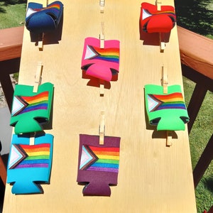 May include: A wooden display board with six colorful can coolers featuring a rainbow and a pink triangle design. The coolers are held in place with wooden clothespins.