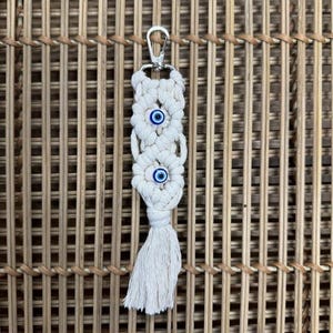 Puede incluir: Llavero de macram&eacute; blanco con un llavero plateado y tres dijes de ojo azul.