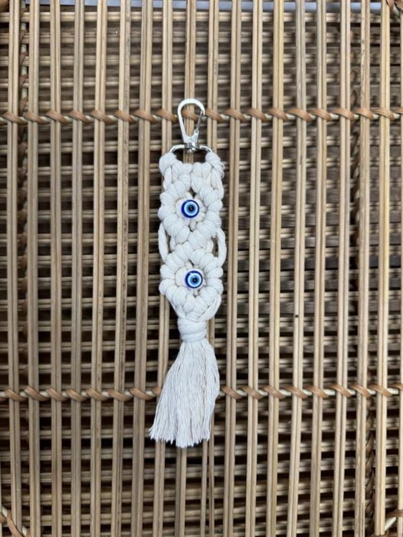Puede incluir: Llavero de macram&eacute; blanco con un llavero plateado y tres dijes de ojo azul.