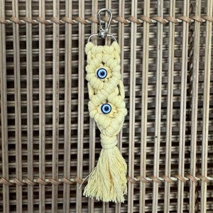 Puede incluir: Un llavero de macram&eacute; amarillo con tres amuletos de ojo azul. El llavero tiene un anillo de metal plateado.