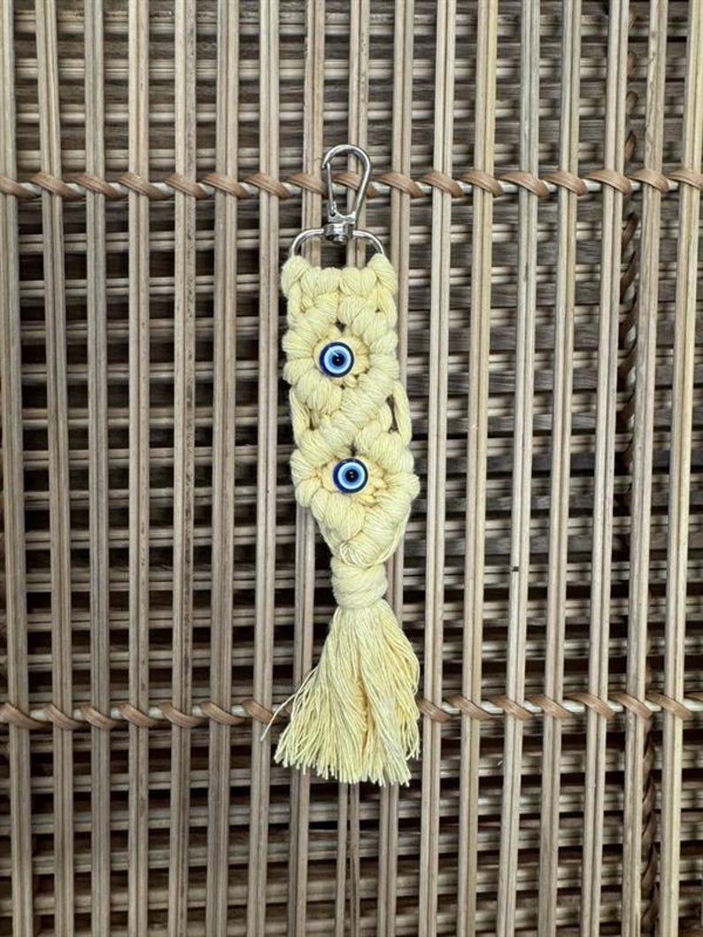 Puede incluir: Un llavero de macram&eacute; amarillo con tres amuletos de ojo azul. El llavero tiene un anillo de metal plateado.