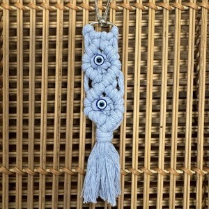 Puede incluir: Un llavero de macram&eacute; azul claro con tres amuletos de ojo azul. El llavero tiene un anillo de metal plateado y una borla.