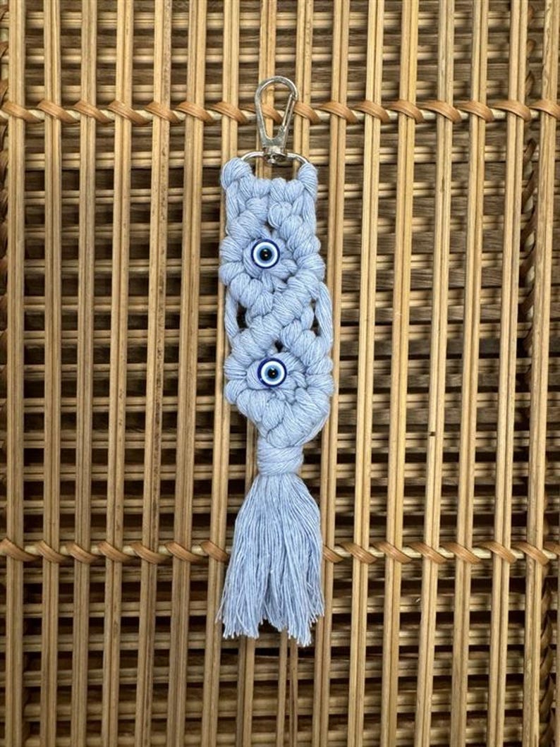 Puede incluir: Un llavero de macram&eacute; azul claro con tres amuletos de ojo azul. El llavero tiene un anillo de metal plateado y una borla.