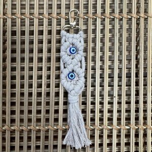 Puede incluir: Un llavero de macram&eacute; blanco con un borla y tres abalorios de ojo azul. El llavero est&aacute; unido a un anillo de metal dorado.