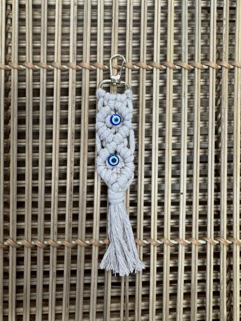 Puede incluir: Un llavero de macram&eacute; blanco con un borla y tres abalorios de ojo azul. El llavero est&aacute; unido a un anillo de metal dorado.