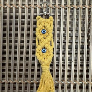 Puede incluir: Un llavero de macram&eacute; amarillo con tres dijes de ojo azul y una borla.