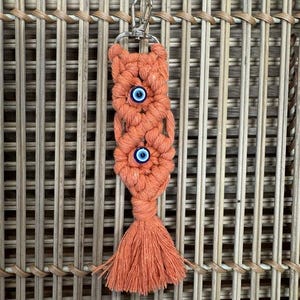 Puede incluir: Un llavero de macram&eacute; de color coral con dos amuletos de ojo azul y una borla.