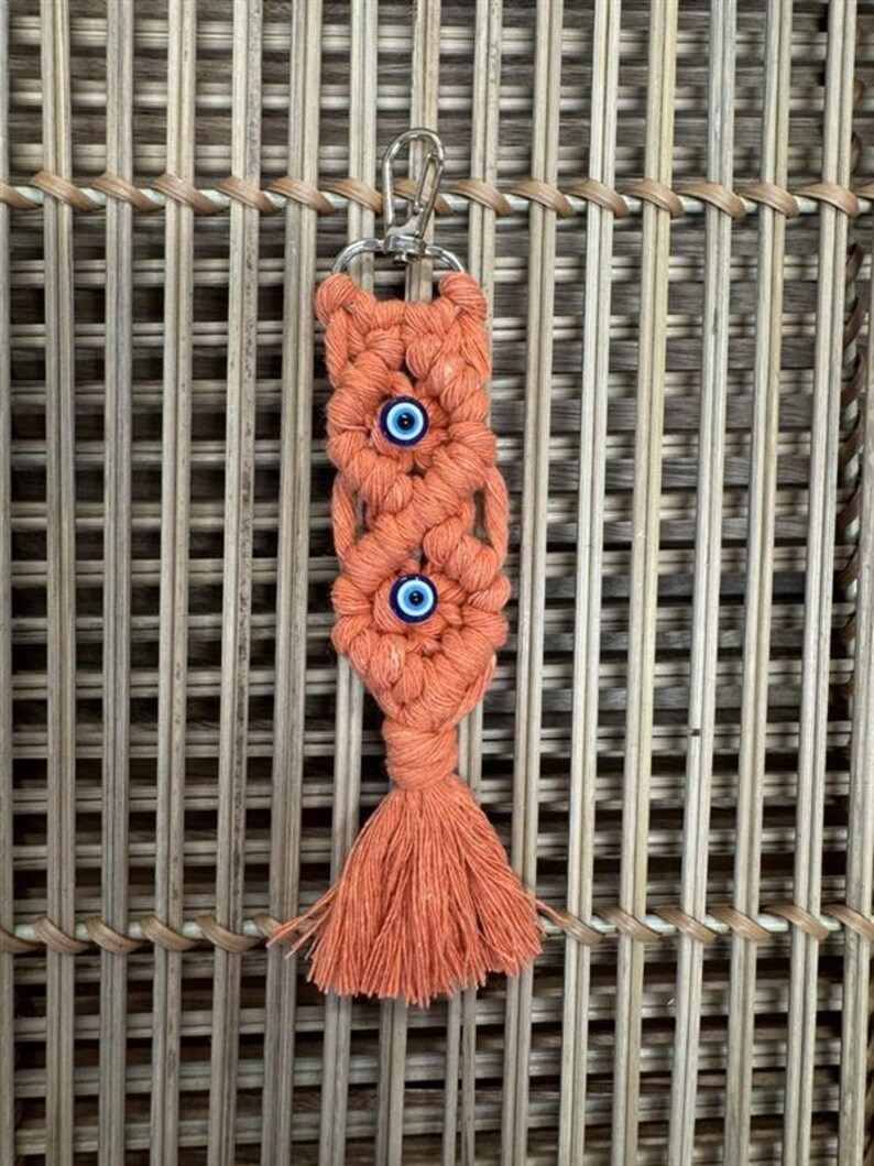 Puede incluir: Un llavero de macram&eacute; de color coral con dos amuletos de ojo azul y una borla.