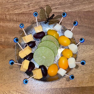 Op de afbeelding: Een zilveren schaal met een vlinderontwerp bevat een verscheidenheid aan snacks op spiesjes. De spiesjes bevatten kaasblokjes, olijven, kumquats en limoenpartjes. De schaal is versierd met blauwe en witte boze oogcharms.