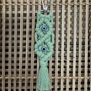Puede incluir: Un llavero de macram&eacute; verde con un anillo de llave plateado y dos amuletos de ojo azul. El llavero tiene un borla en la parte inferior.
