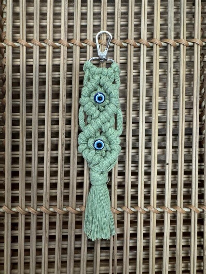 Puede incluir: Un llavero de macram&eacute; verde con un anillo de llave plateado y dos amuletos de ojo azul. El llavero tiene un borla en la parte inferior.