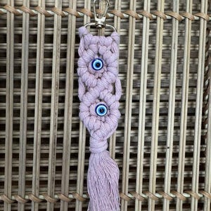 Puede incluir: Un llavero de macram&eacute; rosa con un borla y dos dijes de ojo azul.