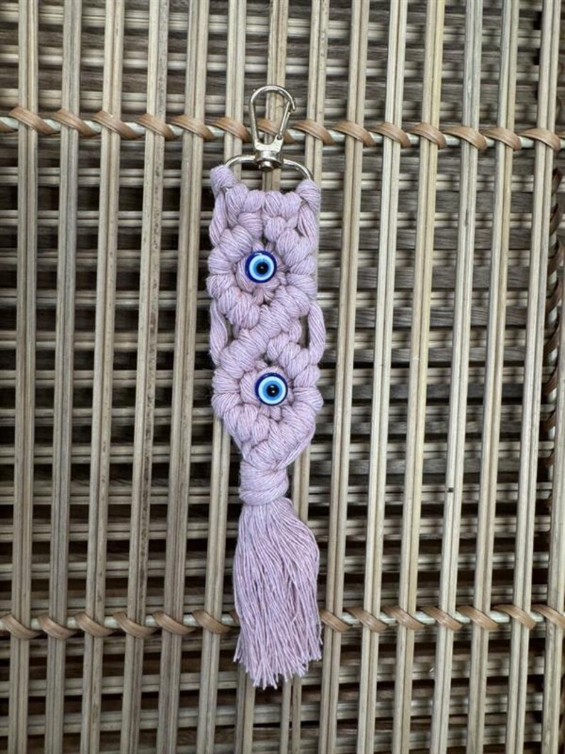 Puede incluir: Un llavero de macram&eacute; rosa con un borla y dos dijes de ojo azul.