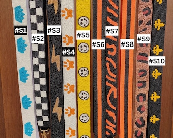 May include: A collection of ten beaded purse straps with various designs. The straps feature patterns such as crowns, checkered squares, lightning bolts, paw prints, smiley faces, and leopard print. Each strap has a gold-colored clasp and is labeled with a number.
