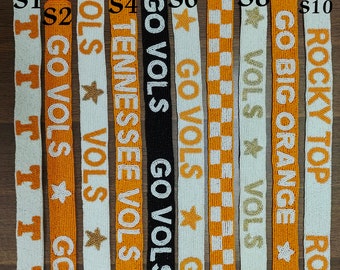 May include: A selection of beaded straps in various designs. The straps feature white, orange, and black beads with text like "GO VOLS", "TENNESSEE", and "ROCKY TOP". Some straps have star accents and checkerboard patterns.