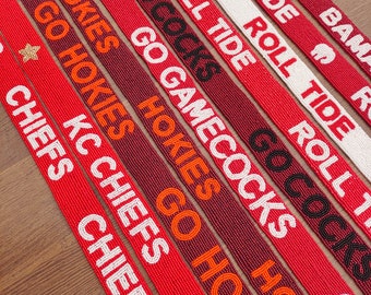 May include: Close-up of several beaded lanyards in red, white, and maroon. The lanyards feature team names and phrases like "Roll Tide," "Go Cocks," and "KC Chiefs" in various colors. Some lanyards have a star embellishment.